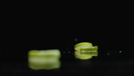 Slow-motion.-Sliced-​​lime-rings-fall-with-splashes-of-water-on-the-glass-on-a-dark-background.-Cut-into-slices